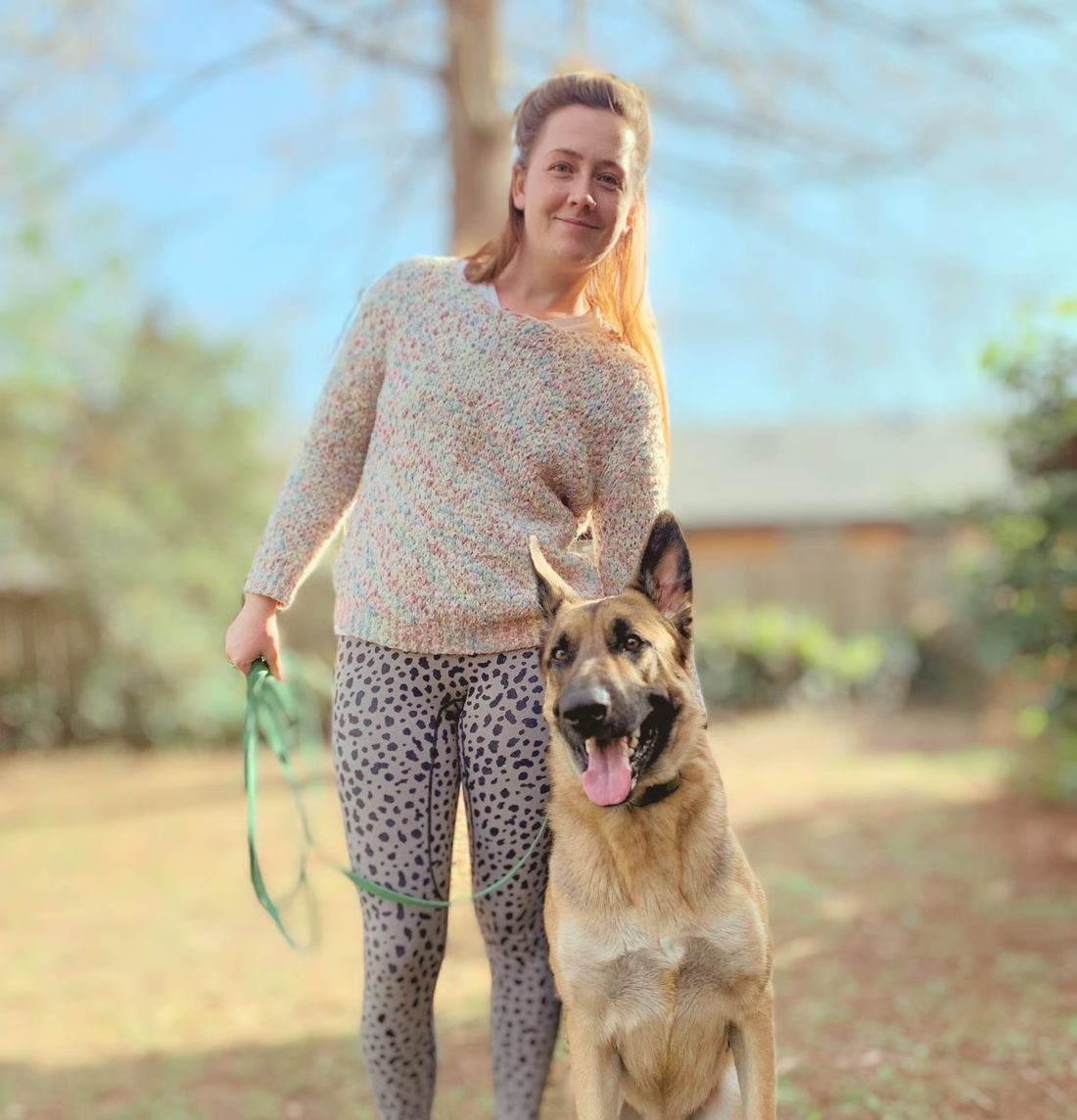 Marta standing outdoors holding a leash beside a German shepherd sitting calmly at her side